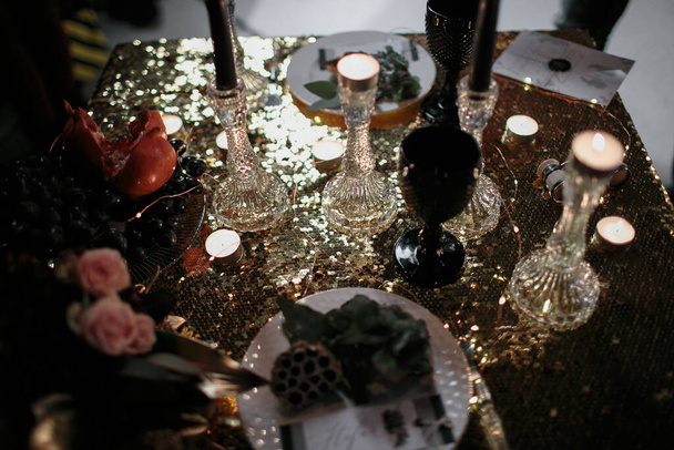 Medium sharp front view of decorated wooden table with burning black candles with pomegranate, grape, binoculars and ceremonial serving on tables at luxury wedding reception in restaurant in twilight.  - Photo, image