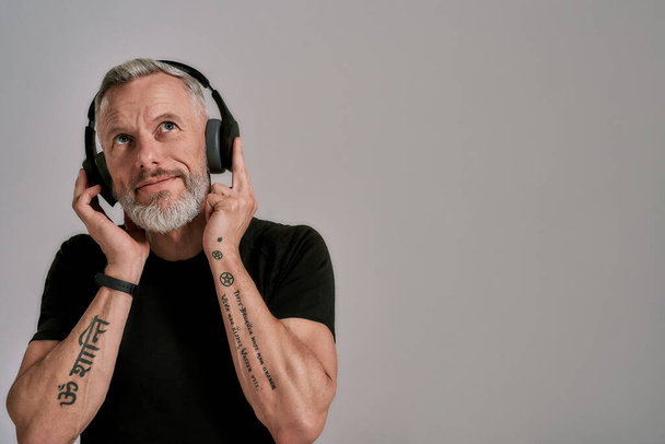 Dreaming. Middle aged muscular man in black t shirt and headphones looking up while listening to music, posing in studio over grey background - Zdjęcie, obraz
