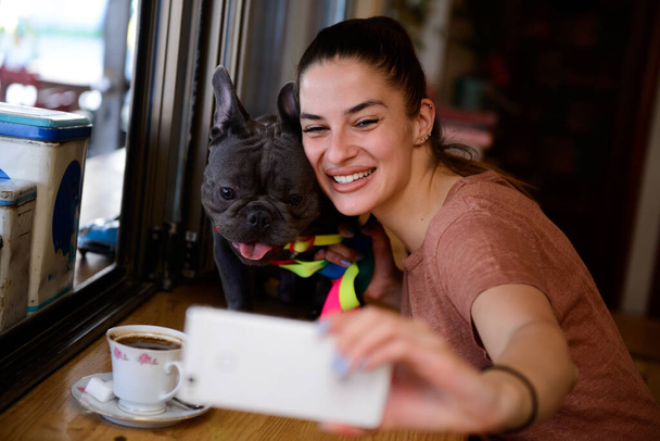 Nuori tyttö nauttii vapaa-ajastaan hänen ihastuttavan Blue French Bulldogin kanssa ja ottaa selfiet pienen, viihtyisän, maalaismaisen lemmikkiystävällisen kahvilan sisällä.. - Valokuva, kuva