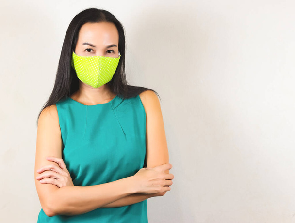 Krásná asijská žena v puntíkové zelené hygienické masce a zeleném tričku bez rukávů, přes paže a při pohledu na kameru.Móda a ochrana před covid-19 konceptem. - Fotografie, Obrázek