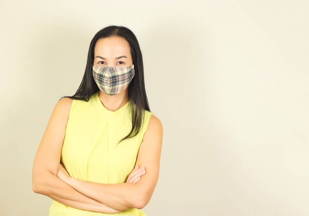 Belle femme asiatique portant un masque hygiénique à carreaux jaunes et bleus et un t-shirt jaune sans manches, croisant les bras et regardant la caméra.Mode et protection contre le concept covid-19. - Photo, image
