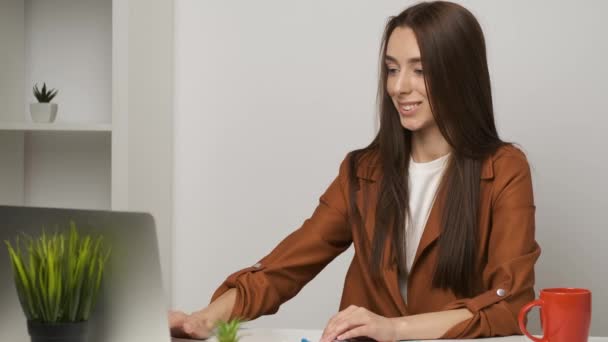 Female brunette accountant sitting at the desk working on a laptop and smiling sincerely - Кадры, видео