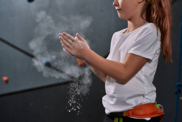 Cropped shot of sportive kid, girl applying white dust of magnesia, chalk on hands before climbing wall in bouldering center. Concept of sport life and rock climbing - Foto, immagini