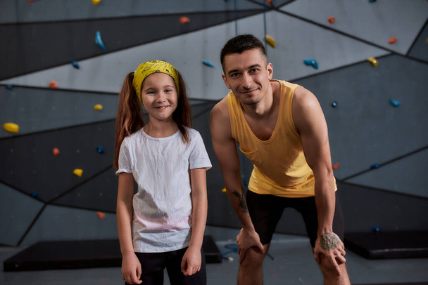 Young male instructor and active little girl looking at camera, standing against artificial training climbing wall. Concept of sport life and rock climbing - Фото, изображение