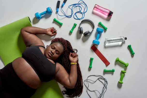 Better change. Top view of tired plump, plus size african american woman in sportswear lying on yoga mat after workout with sports, fitness equipment and headphones near her over grey background - Fotoğraf, Görsel