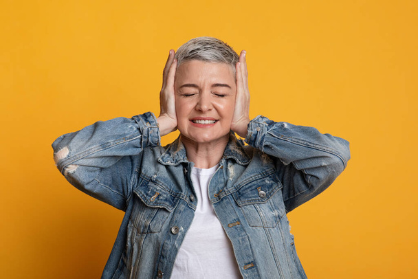 Geïrriteerde vrouw van middelbare leeftijd bedekt oortjes in stress, staande over gele achtergrond - Foto, afbeelding