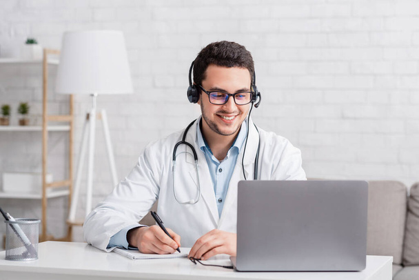 Lächelnder Arzt im weißen Kittel mit Kopfhörer, macht Notizen in Notizbuch-Optik im Laptop - Foto, Bild
