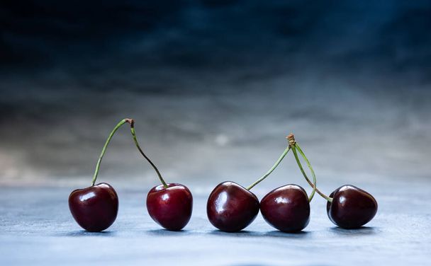 Close-up of ripe juicy dark red and black sweet cherries. Five sweet cherries in a row on blue surface. Horizontal background with copy space. Healthy eating and dieting concept. Selective focus. - Foto, Imagem