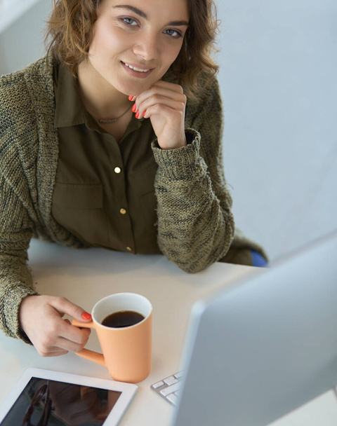 Attractive smiling woman sitting at office desk, holding a cup of coffee, she is relaxing and looking away - Фото, изображение