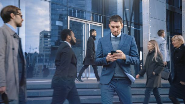 Uomo d'affari caucasico in giacca e cravatta sta utilizzando uno smartphone su una strada nel centro della città. Altre persone dell'ufficio camminano oltre. Sorride e sembra di successo. Sta navigando sul Web sul suo dispositivo. - Foto, immagini