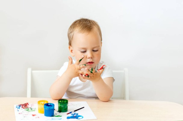 kind jongen 2 jaar oud zitten aan een tafel op een witte geïsoleerde achtergrond en verf, vroege ontwikkeling, plaats voor tekst - Foto, afbeelding