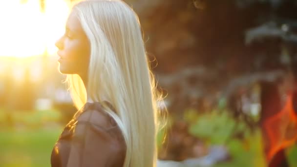 Caucasian girl gracefully goes towards the setting sun past green trees in the park. Long blond fluffy hair flutters from behind, the teenager turns around, looks confidently at the camera and smiles. - Video