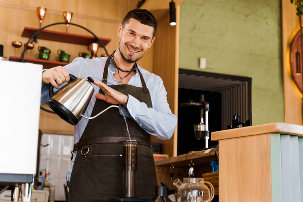 Handsome barista pours hot water in aeropress with coffee and look in camera and smile in cafe. Bearded barista works. Alternative making by barista in the cafe. Scandinavian coffee brewing method. - Photo, Image