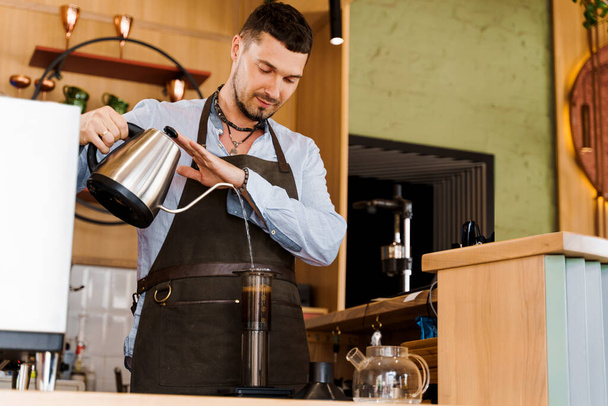 El camarero guapo vierte el agua caliente en aeroprensado con el café en la cafetería. El camarero barbudo trabaja. Fabricación alternativa por barista en el café. Método de elaboración del café escandinavo
. - Foto, imagen