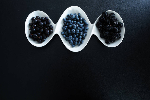 Set of different types of black berries in a white plate on a black table. Stylish seasonal vitamins. - Valokuva, kuva