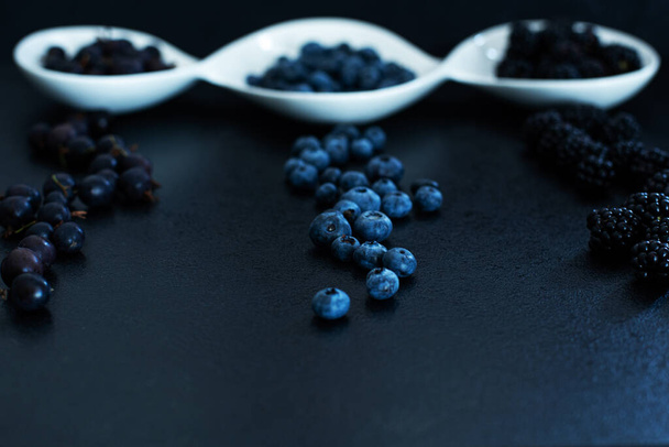 Set of different types of black berries in a white plate on a black table. Stylish seasonal vitamins. - Photo, image
