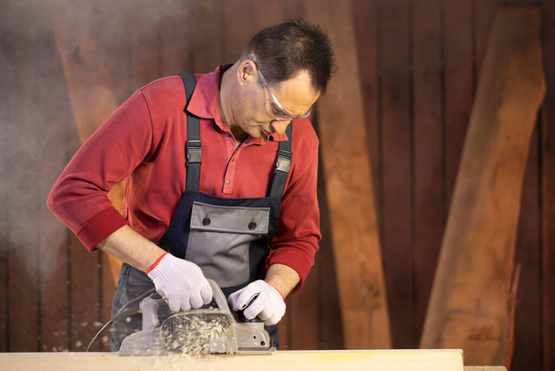 homem de meia-idade artesão em googles trata pedaço de madeira com plaina elétrica
 - Foto, Imagem
