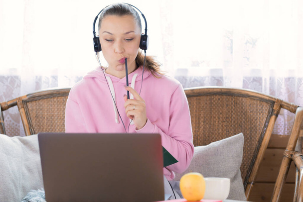 girl in pink hoodie and headphones thoughtfully looking at laptop chewing pencil - Photo, Image