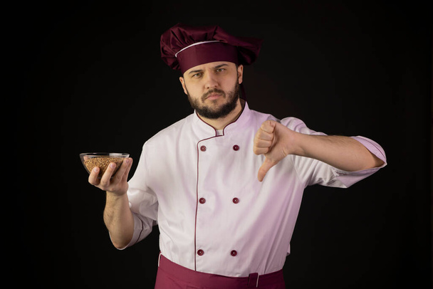 Unsatisfied chef man in uniform holds bowl with buckwheat shows dislike sign - Φωτογραφία, εικόνα