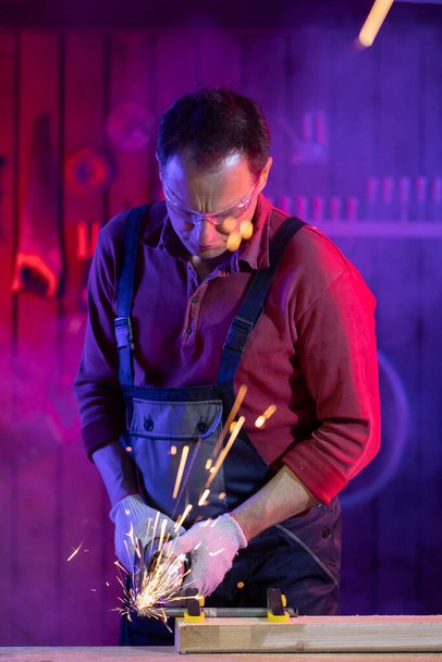 mężczyzna w kombinezonach gogle i rękawice szlifowanie metalu iskry w garażu - Zdjęcie, obraz