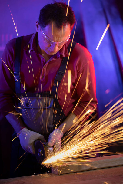 man in jumpsuit goggles and gloves grinding metal with sparks in coloured light - Photo, Image