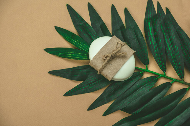 bílá mýdlová tyčinka pokrytá prázdným řemeslným papírem na plochém palmovém listu ležela béžové pozadí - Fotografie, Obrázek