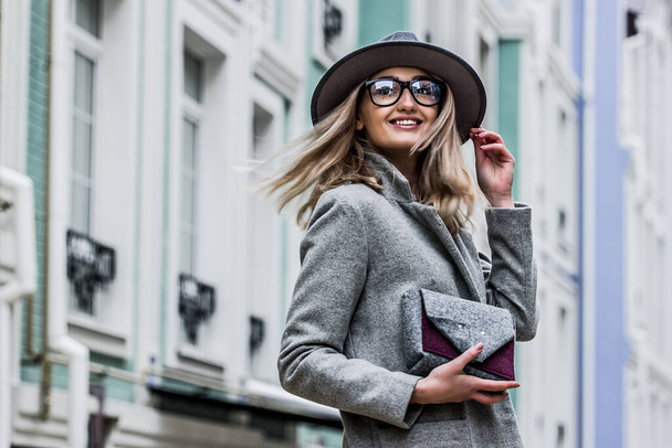 Fashionable hipster girl in eyeglasses and hat looking away and smiling while standing on city street. Portrait young woman in coat with hair in motion outdoors. Woman with handbag. City style, outfit - Fotó, kép