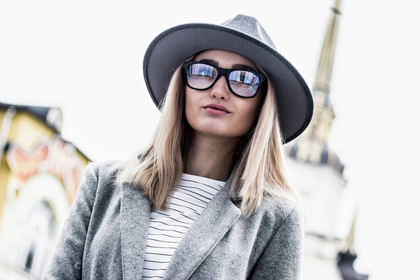 Beautiful, fashionable hipster girl in hat and eyeglasses posing to camera while standing outdoors. Portrait stylish young woman in coat walking on city street. Modern look for city girl. Urban outfit - Foto, afbeelding