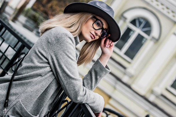Portrait of affectionate stylish hipster girl touching hat while standing on balcony outdoors. Young beautiful woman in eyeglasses posing on city street. Thoughtful woman dreaming on city street - Фото, изображение