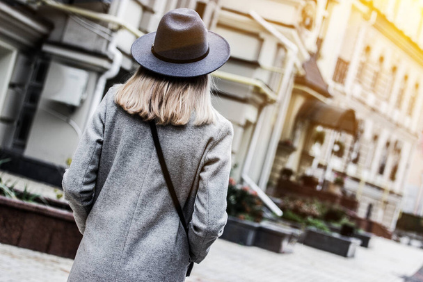 Back view of young stylish blonde woman in hat and coat walking on city streets. Attractive fashionable hipster girl standing outdoors. Modern style of city fashion. Urban outfit for young people - Fotó, kép