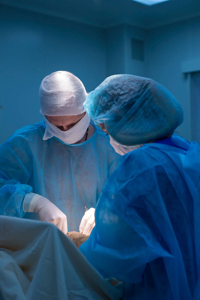 A gyermeksebészek urológiai műtéteket végeznek. Egy férfi és egy nő maszkban és egy kék, steril kabátban a műtőben. Egy csapat sebész végzi a műtétet. A lágyéksérv, hereödéma, herebetegség, varicocel kezelése - Fotó, kép