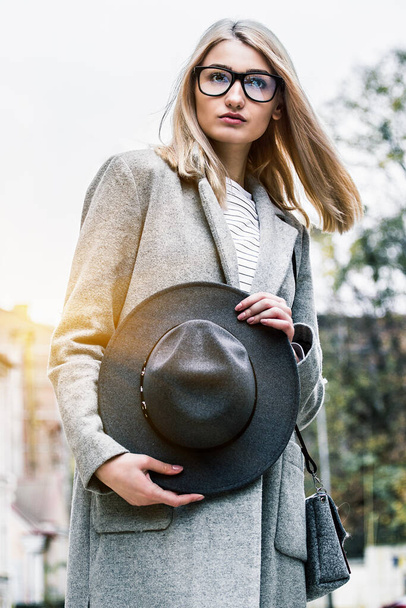 Portrait of charismatic young woman looking to camera and holding hat in hands while standing outdoors. Fashionable and pretty hipster girl in coat posing outdoors. Modern look for stylish city girl - 写真・画像