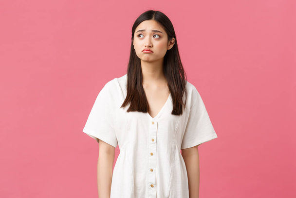 Beauty, people emotions and summer leisure concept. Sad and gloomy asian lonely girl looking upset and depressed, pouting and frowning as looking upper left corner, pink background - Photo, Image