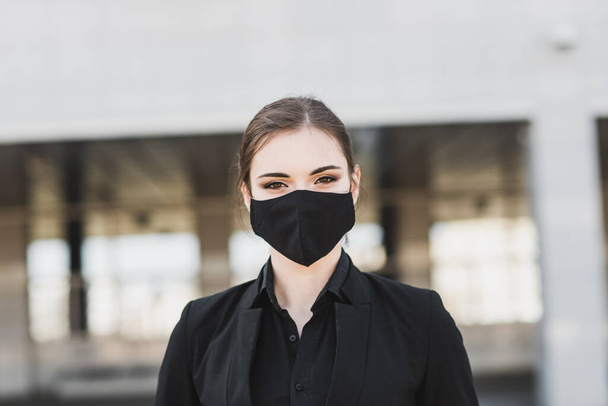Siyah takım elbiseli, siyah maskeli ve eldivenli güzel bir iş kadını şehirde karantina ve tecrit altında. Salgın COVID 19. Seçici odaklı resim - Fotoğraf, Görsel