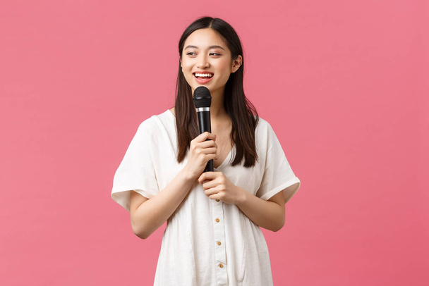 Vapaa-ajan, ihmisten tunteita ja elämäntapa käsite. Iloinen hymyilevä aasialainen tyttö karaokessa, nauttia viikonloppuisin, laulaa laulu mikrofonissa, suorittaa stand-up, seisova vaaleanpunainen tausta - Valokuva, kuva