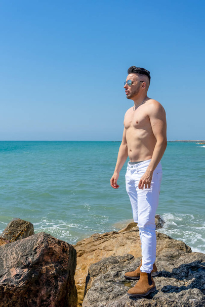 Νεαρός άνδρας με λευκό παντελόνι και μοντέρνο στυλ ποζάρει στην παραλία της Μεσογείου στη Μπουριάνα. Έννοια μόδας - Φωτογραφία, εικόνα