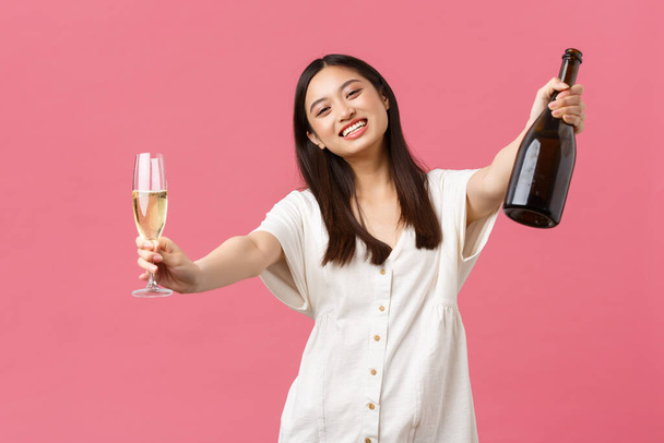 Oslava, svátky a zábavný koncept. Šťastná usměvavá asijská dívka hostitel akce, drží láhev šampaňské a sklo a natahuje ruce pro objímání hosta, stojící růžové pozadí - Fotografie, Obrázek
