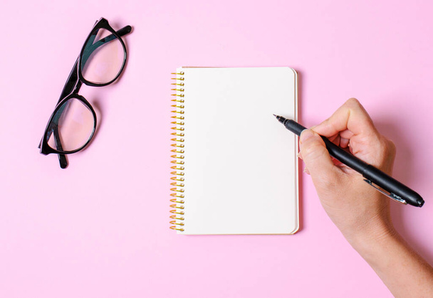 Notebook with clean sheets for records, hand holds pen, office glasses on a pink background. Flat lay - Photo, Image