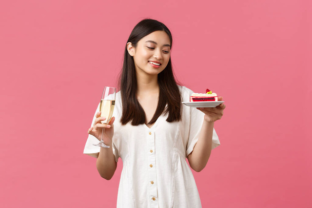 Feiern, Feiertage und Spaßkonzept. Verträumt schöne Frau feiert ihren Geburtstag mit Glas Champagner und B-Day-Kuchen, lächelt unbeschwert zum Dessert, steht rosa Hintergrund - Foto, Bild