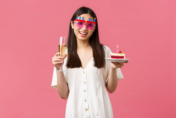 Celebration, party holidays and fun concept. Cheerful happy asian woman celebrating birthday in funny sunglasses, holding glass champagne and b-day cake with lit candle to make wish - Фото, зображення