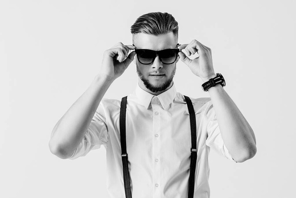 Black and white shot of stylish and elegant man in shirt with suspenders wearing sunglasses while standing against white background. Fashionable man in classical clothing. Men's fashion and style - Photo, Image