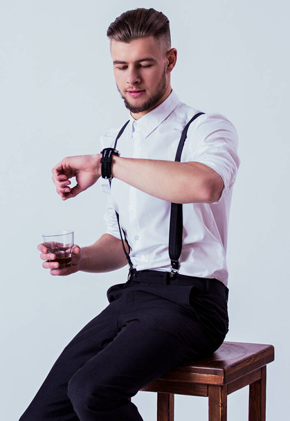 Studio shot elegant and confident man in classical wear checking time on watches and drinking whiskey while sitting on chair or stool against gray background. Fashionable man in white shirt,suspenders - Foto, Imagem