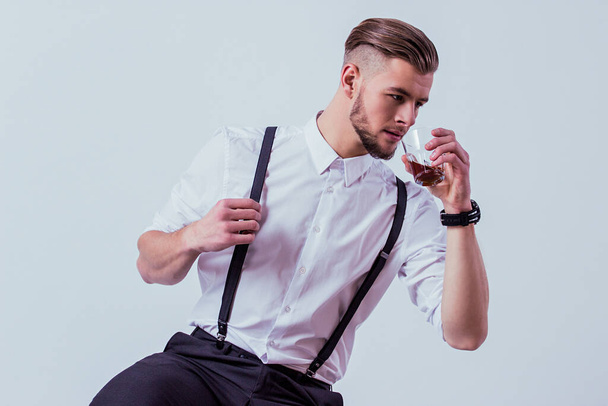 Studio shot of handsome bearded man in white shirt and suspenders drinking alcohol while sitting against gray background. Elegant man in classical wear with glass of whiskey in hand. Style, fashion - Foto, Bild