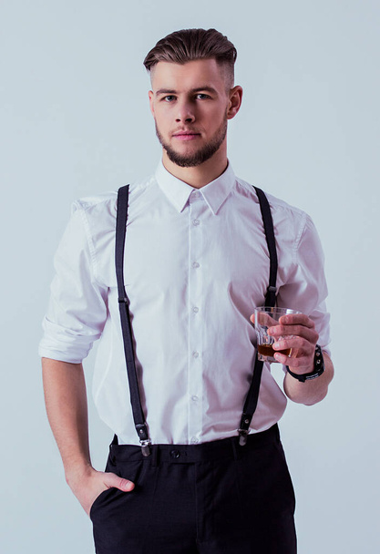 Confident and elegant man in classical wear keeping hand in pocket and looking to camera while drinking whiskey or scotch against gray background. Men with glass of alcohol. Masculinity and charisma - Fotoğraf, Görsel