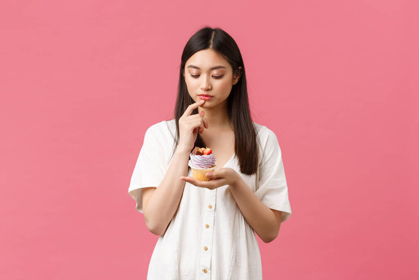 Jídlo, kavárna a restaurace, letní životní styl. Promyšlený roztomilý váhavý asijské žena při pohledu na cupcake, přemýšlet o kaloriích jako pobyt na dietě, lákavé jíst lahodný dezert - Fotografie, Obrázek
