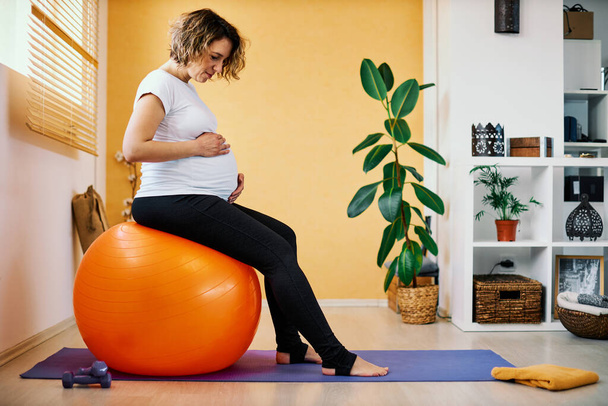 Femme enceinte d'âge moyen assis sur pilates balle et toucher le ventre. - Photo, image