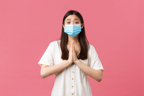 Covid-19, társadalmi távolság, vírus és életmód koncepció. Reményteli, buta ázsiai lány segítségért könyörög, mondja, kérem, és fogja meg a kezét könyörgésben, orvosi maszkban és nyári ruhában rózsaszín háttér felett - Fotó, kép