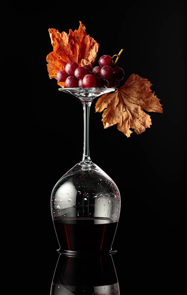 Inverted wine glass with red wine on a black reflective background. Wine with grapes and dried vine leaves. Concept of winemaking. - Fotó, kép