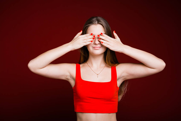 A young girl with long hair and a red T-shirt with her hands closed her eyes so as not to see what is happening around on a burgundy background - Foto, Imagen