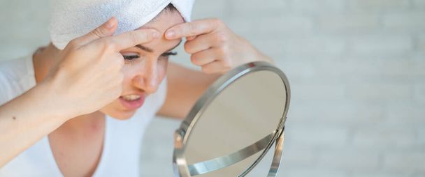 An upset woman looks in the mirror and squeezes a pimple on her forehead. A girl with hormonal problems cleanses her face after a shower. Teen acne. Himself a dermatologist. Problem skin. - Фото, изображение